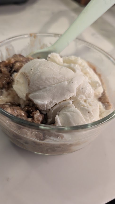 A glass bowl of warm apple crumble topped with a scoop of vanilla ice cream