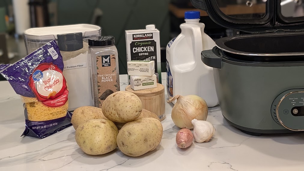 Potatoes, shredded cheddar, milk, broth, onion, and garlic on a kitchen counter next to a Ninja multicooker